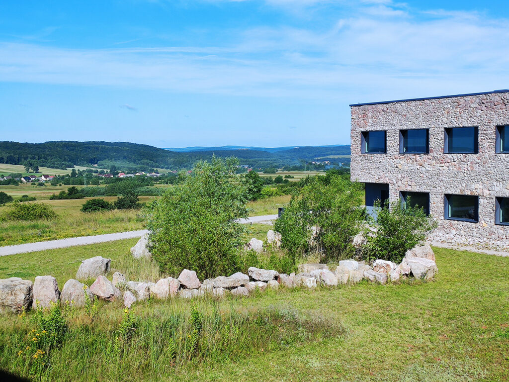 Modern stone building of ECGC with large windows bordering a lush green landscape and a stone fence. Background features rolling hills under a blue sky.