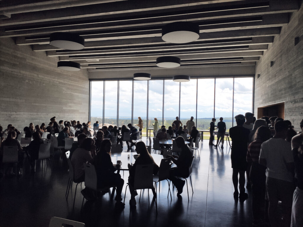 A spacious cafeteria with high ceilings and large windows showing a scenic view. Groups of people sit or stand, engaged in conversations. Calm atmosphere.
