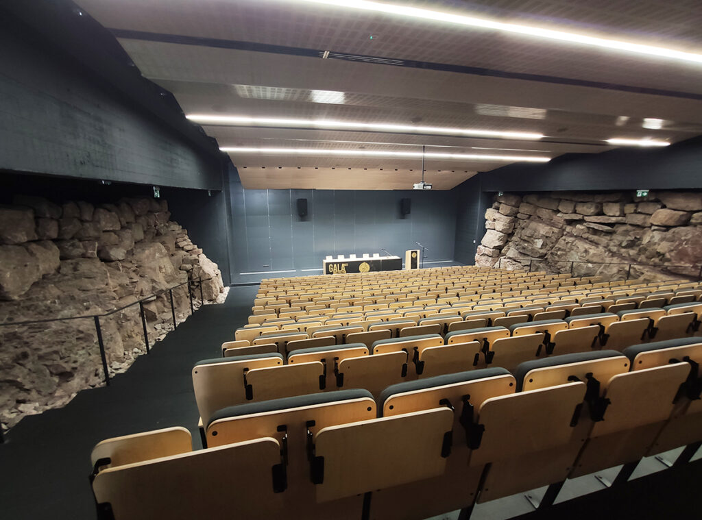 A modern lecture hall tiered seating and stone walls creating a natural ambiance. The room is empty, well-lit, and conveys a serene atmosphere.